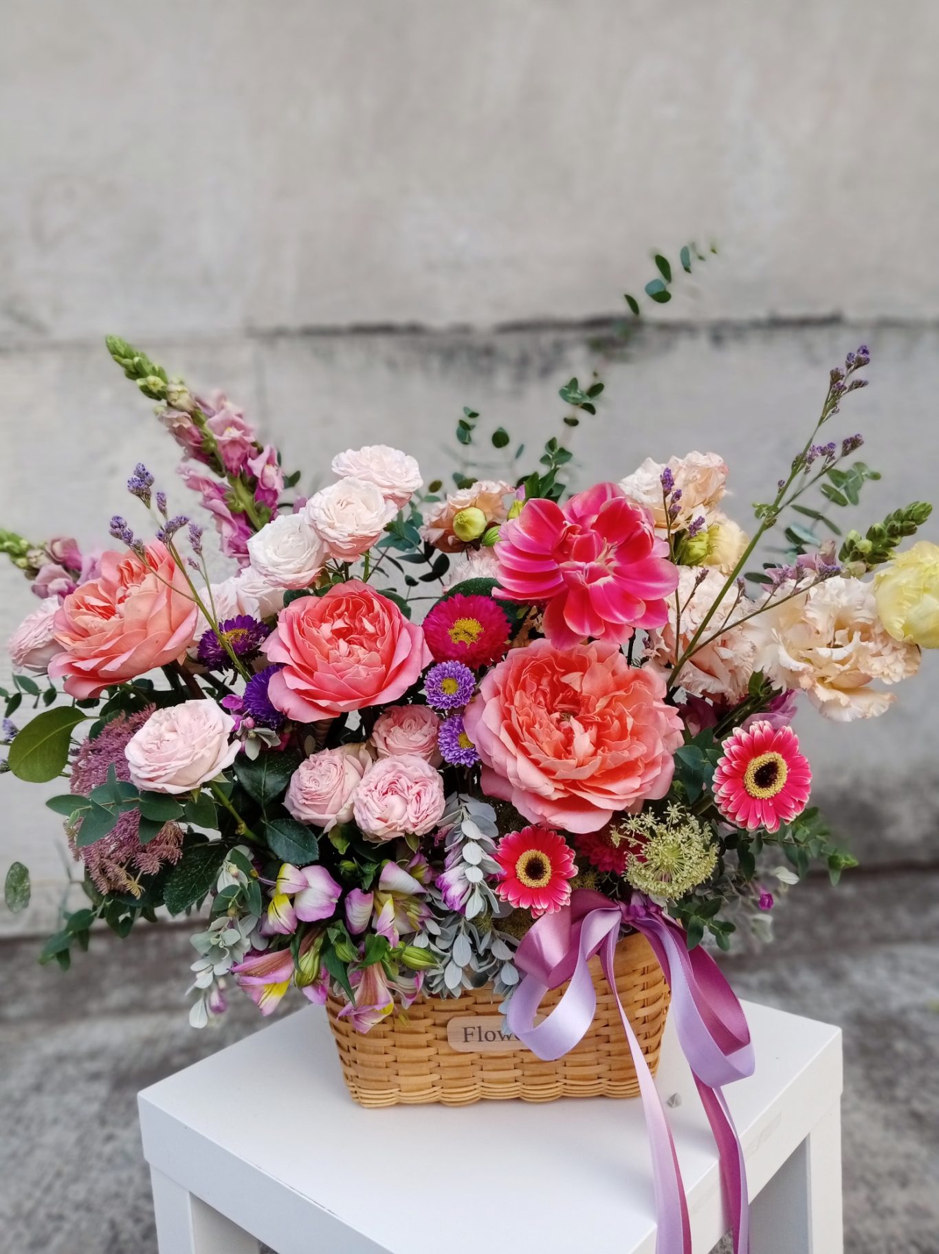 Ramo de flores variadas en una canasta, con tonos rosa y morado decorados con cinta.